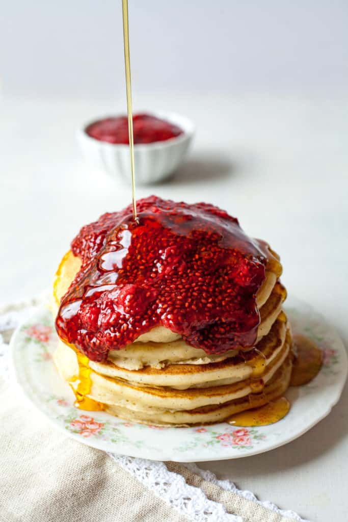 syrup being poured on a stack of pancakes topped with chia jam.
